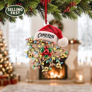 May include: A Christmas ornament shaped like an atom with a red Santa hat on top. The atom features a central cluster of red and green spheres surrounded by gold orbital rings decorated with small, multi-colored Christmas lights. The name CAMERON is printed in black on the white brim of the hat.