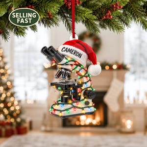 Könnte beinhalten: Ein Mikroskop-Weihnachtsanhänger mit einer roten Weihnachtsmütze, auf der der Name CAMERON in Schwarz gedruckt ist. Das Mikroskop ist mit einer bunten Lichterkette umwickelt und hängt an einem roten Band vor einem festlichen Hintergrund.