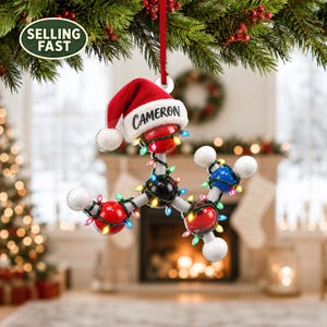 May include: A molecular structure Christmas ornament featuring red, black, and blue spheres connected by grey rods. The ornament is wrapped in miniature multi-coloured string lights and topped with a red and white Santa hat labelled CAMERON.