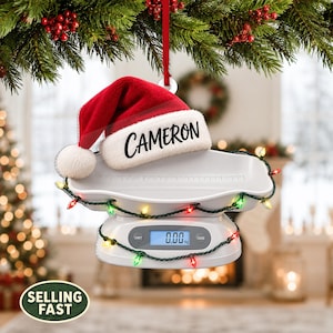 Può includere: Un ornamento a forma di bilancia digitale per neonati bianca con un cappello da Babbo Natale rosso e bianco con il nome CAMERON stampato in nero sul bordo. La bilancia è avvolta da una catena di luci natalizie multicolori e segna 0,00 kg.