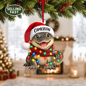 Könnte beinhalten: Ein Weihnachtsschmuck in Form eines Baby-Alligators, der eine rote Weihnachtsmütze mit dem Namen CAMERON auf dem weißen Rand trägt. Der Alligator ist in einen roten Schal und eine Lichterkette mit bunten, leuchtenden Lichtern gewickelt und hängt an einem roten Band.
