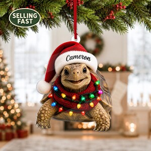 以下が含まれることがあります： クリスマスツリー用の平らな吊り下げ式オーナメント。サンタの帽子をかぶったカメが描かれており、帽子の縁にはCameronという名前が印刷されています。カメは赤いニットのマフラーを巻いており、そのマフラーには色とりどりのクリスマスライトが飾られています。