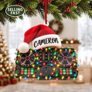 Puede incluir: Un adorno de controlador de DJ con un gorro de Papá Noel rojo con el nombre CAMERON impreso en negro en el borde blanco. El controlador está envuelto en una cadena de luces navideñas multicolores y cuelga de una cinta roja.