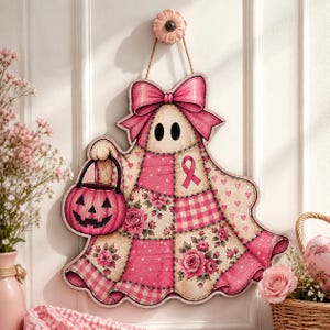 May include: A decorative wall hanging shaped like a ghost, featuring a patchwork design in shades of pink and cream with floral and gingham patterns. The ghost has a large pink bow, a pink ribbon symbol, and holds a pink jack-o'-lantern bucket.