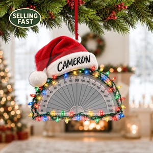 May include: A semi-circular protractor Christmas ornament featuring a red Santa hat with the name CAMERON printed in black. The protractor is decorated with a string of colourful miniature Christmas lights along its curved edge.