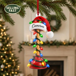 May include: A Christmas tree ornament shaped like a red rubber plunger with a wooden handle. The plunger is wrapped in a string of multicolored LED lights and topped with a red and white Santa hat featuring the name Cameron printed in black script.