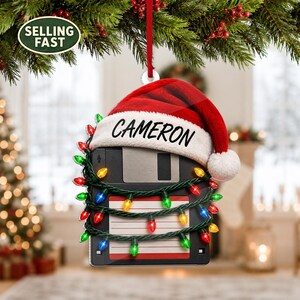 May include: A black floppy disk Christmas ornament featuring a red and white Santa hat with the name CAMERON printed on the brim. The disk is wrapped in a string of multicoloured Christmas lights with red, green, blue, and yellow bulbs.