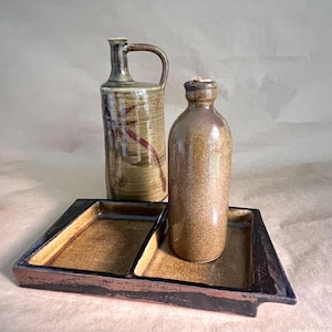 May include: A ceramic set consisting of a rectangular tray with two compartments, a tall handled pitcher with brown and tan glaze, and a smaller cylindrical bottle with a speckled brown finish. The tray features a dark brown rim and base.