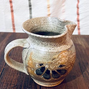 May include: A speckled ceramic creamer jug with a rounded body and a curved handle. The exterior features a beige and grey speckled glaze with a prominent embossed floral design in shades of brown and dark grey on the front of the vessel.