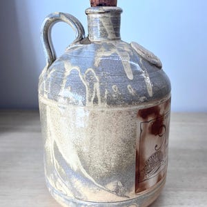 May include: A ceramic jug with a cork stopper, featuring a mottled grey and beige glaze with vertical drip patterns. A brown rectangular label on the side displays the text Old Clare, with a circular logo and the words Country Store printed below.