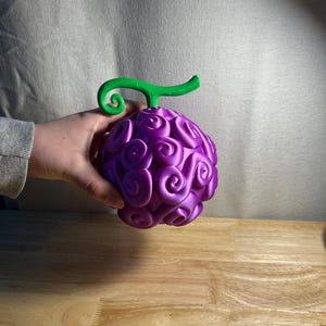 May include: A purple, spherical object with a green stem, held by a hand. The object has a textured surface with swirling patterns. The stem is curved and has a spiral design. The object is resting on a wooden surface.