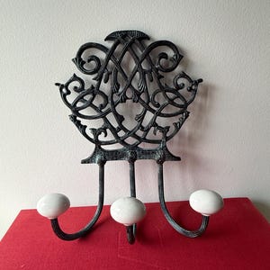 May include: A wall-mounted coat rack featuring a dark, weathered metal frame with intricate, scrolling openwork patterns. The rack includes three curved metal hooks, each tipped with a smooth, round, white ceramic knob for hanging items.