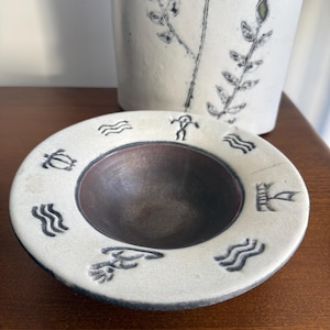 May include: A ceramic bowl with a wide, cream-coloured rim featuring dark, etched symbols including waves, a turtle, and a figure. The interior of the bowl is a deep, dark brown. Behind it stands a tall, cylindrical cream-coloured ceramic vase.