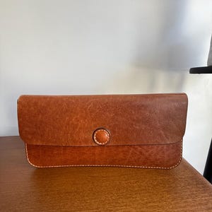 May include: A rectangular brown leather clutch wallet featuring a smooth flap closure secured by a single circular leather button. The wallet is constructed with visible light-coloured stitching along the bottom edge and around the central button.