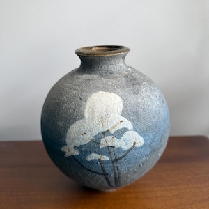 May include: A spherical ceramic vase with a textured, dark grey and blue mottled finish. The front features a hand-painted white floral design with thin black stems. The vase has a narrow, circular opening with a smooth, brown-toned rim.
