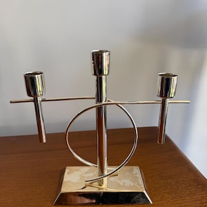May include: A silver-toned metal candelabra featuring three candle holders arranged on a horizontal bar. The structure includes a central vertical post, a circular ring element, and a rectangular base, all finished with a polished, reflective surface.