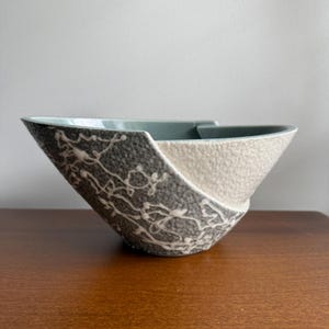 May include: A ceramic bowl with an asymmetrical, slanted rim. The exterior features a textured surface divided into two sections: one side is dark grey with white, vine-like raised patterns, and the other side is a solid, off-white textured finish.