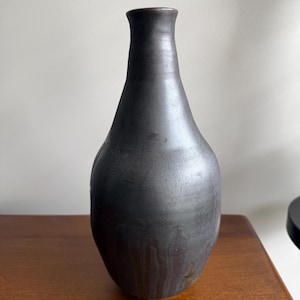 May include: A tall, slender ceramic vase with a matte, dark charcoal-grey finish. The vessel features a narrow neck that gradually widens into a rounded, bulbous body, showcasing subtle, vertical drip-like textures in the glaze.