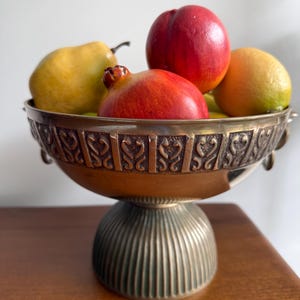 May include: A metallic pedestal bowl with a wide, fluted base and a decorative band featuring repeating heart-like motifs. The bowl is filled with a variety of artificial fruits, including a yellow pear, a red pomegranate, a red nectarine, and a lemon.