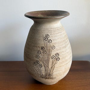 May include: A beige ceramic vase with a flared rim and a dark brown interior. The exterior features a textured, speckled surface with an incised, dark-toned botanical design of thin stems and small circular flower heads on the front.