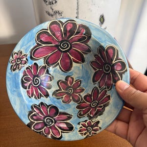 May include: A round ceramic bowl featuring a light blue painted background with several hand-painted dark pink flowers. Each flower has thick black outlines and a spiral centre. The bowl has a smooth, glossy finish and a slightly irregular, organic shape.