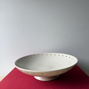 May include: A shallow, off-white ceramic bowl with a pedestal base and a series of small, evenly spaced circular perforations along the rim. The bowl is resting on a red book titled LOVE: A Celebration in Art and Literature.
