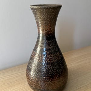 May include: A ceramic vase with a narrow neck and a wide, rounded base. The surface features a textured, mottled glaze in shades of dark brown, bronze, and black, creating a gradient effect that transitions from a lighter top to a darker body.