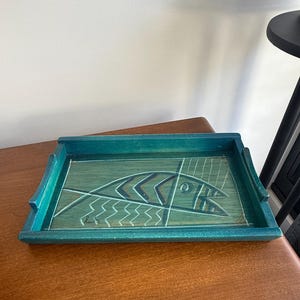 May include: A rectangular teal wooden tray featuring a stylised fish illustration on its base. The fish design is composed of white, black, and brown lines with a geometric pattern. The tray has raised edges and a signature in the bottom left corner.