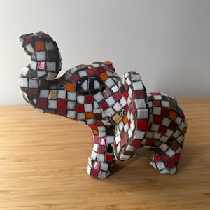 May include: A decorative elephant figurine covered in a mosaic pattern of small square tiles. The tiles are coloured in shades of red, white, orange, and dark brown, creating a checkered effect across the entire surface of the animal sculpture.