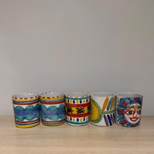 May include: Five cylindrical ceramic cups with hand-painted designs. The cups feature patterns of blue, yellow, and red, with one cup depicting a smiling sun face. The cups are displayed on a wooden surface.