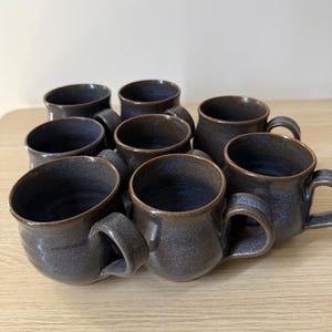 May include: A collection of seven ceramic mugs with a dark, speckled blue and charcoal glaze. Each mug features a rounded body, a wide circular rim, and a sturdy side handle. The finish has a subtle, textured appearance with variations in colour.