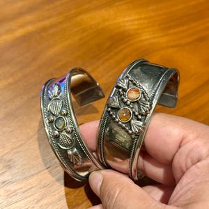 May include: Two silver-toned cuff bracelets featuring intricate metalwork and inlaid stones. One bracelet displays a single central stone, while the other features two stacked stones. Both pieces have decorative leaf-like patterns and rope-style borders.