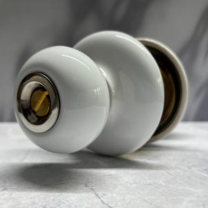 May include: A white ceramic door knob with a smooth, glossy finish and a rounded, spherical shape. The center of the knob features a circular metallic silver insert with a gold-toned slotted screw head visible in the middle of the mechanism.