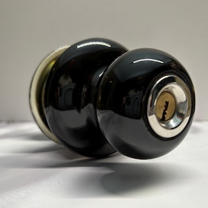 May include: A glossy black spherical door knob featuring a central silver-toned keyhole escutcheon. The knob has a smooth, reflective surface and is attached to a circular base plate with a metallic finish visible at the rear of the assembly.