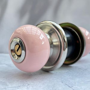 Pode incluir: Um puxador de porta redondo em cerâmica rosa claro com uma base de metal prateado polido e um mecanismo de fechadura em latão ao centro. O puxador apresenta um acabamento liso e brilhante, forma esférica e uma placa de montagem a condizer.