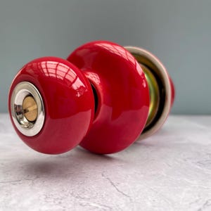 Può includere: Un pomello per porta sferico di colore rosso brillante con una finitura lucida. La parte anteriore presenta una cornice metallica circolare color argento che circonda una fessura centrale per la chiave color ottone. Il pomello ha una forma liscia.