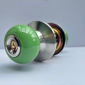 May include: A round, light green ceramic door knob with a polished silver-tone metal keyhole insert at the centre. The knob features a brushed silver-tone circular backplate and a metallic internal mechanism visible behind the main handle.