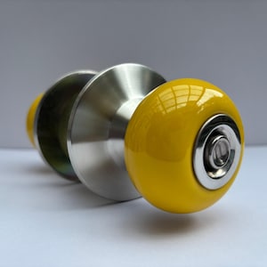 May include: Three cabinet knobs with a bright yellow, rounded face and a silver metal base. The middle knob is silver, and the third knob is yellow. The knobs are likely for a cabinet or drawer.