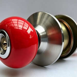 Peut inclure: Un bouton de porte sphérique avec une finition rouge brillante et une serrure métallique centrale. Le bouton est fixé à une plaque circulaire en argent brossé et à un mécanisme interne de couleur laiton, au design épuré et fonctionnel.