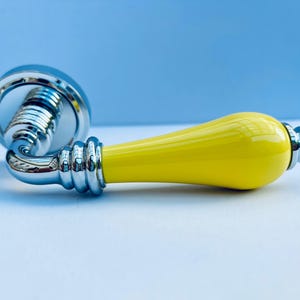 May include: A door handle featuring a bright yellow, smooth, bulbous grip section attached to a polished chrome base. The handle includes a circular mounting plate and a series of decorative, ribbed chrome rings connecting the grip to the base.