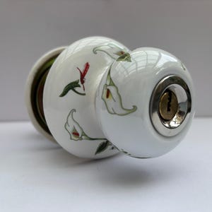 May include: A white ceramic door knob featuring a painted pattern of green stems and white calla lily flowers. The knob has a polished silver-tone metal keyhole insert at the center of the front face and a matching metal base at the rear.