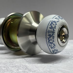 May include: A door knob set featuring two spherical white ceramic handles with a blue floral pattern. The hardware includes a brushed silver metal finish on the circular rosettes and a central keyhole mechanism on the front-facing handle.