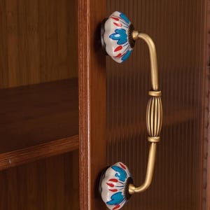 May include: A brass cabinet handle with a curved, ribbed center bar and two decorative ceramic mounts. The mounts feature a white base with a painted pattern of blue petals and red accents, attached to a wooden cabinet door with fluted glass.