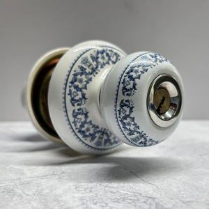 May include: A white ceramic door knob featuring a blue floral pattern. The knob has a round shape with a central silver-toned keyhole insert. The base of the knob is also white with a matching blue floral design, and the internal mechanism is visible.