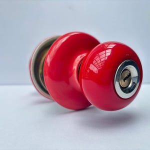 May include: A bright red, spherical door knob with a glossy finish. The front of the knob features a circular silver-toned metal keyhole plate centered on the red surface. The knob has a rounded, smooth profile and a metallic internal mechanism.