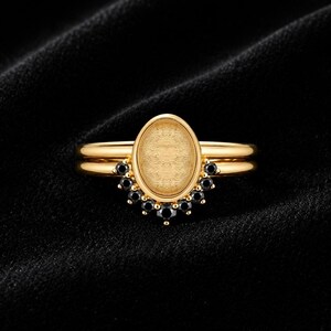 May include: A gold ring featuring a central oval-shaped setting with a textured gold surface. The setting is framed at the base by a semi-circular arrangement of nine small, round, black stones, creating a distinct contrast against the polished gold band.