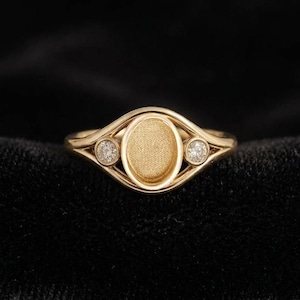 Può includere: Un anello d'oro con un centro ovale e due pietre rotonde più piccole su entrambi i lati. L'anello ha un centro strutturato e un design delicato e fluido. L'anello è posto su uno sfondo nero.