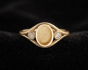 Op de afbeelding: Een gouden ring met een centrale ovale zetting met een matte, getextureerde afwerking. De ringband splitst zich aan beide kanten in twee fijne armen, elk versierd met een kleine, ronde, heldere edelsteen in een ronde zetting.