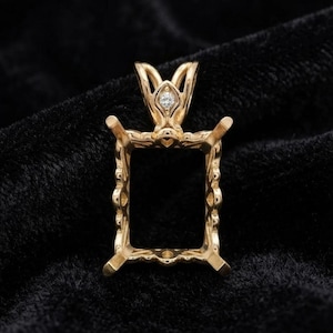 May include: A gold pendant setting featuring a rectangular frame with four prongs for a gemstone. The bail is decorated with a small, round, clear stone set at the top. The metal has a polished finish and intricate, ornate detailing along the edges.
