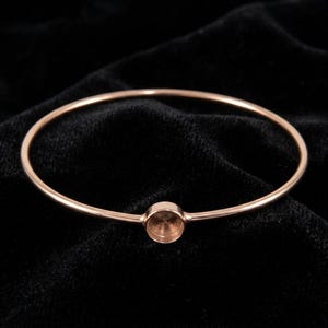 Könnte beinhalten: Ein roségoldenes Armreif-Armband mit einer kreisförmigen Fassung. Das Armband besteht aus dünnem Metall und hat ein schlichtes, elegantes Design. Die Fassung ist für einen kleinen Stein oder Anhänger gedacht.